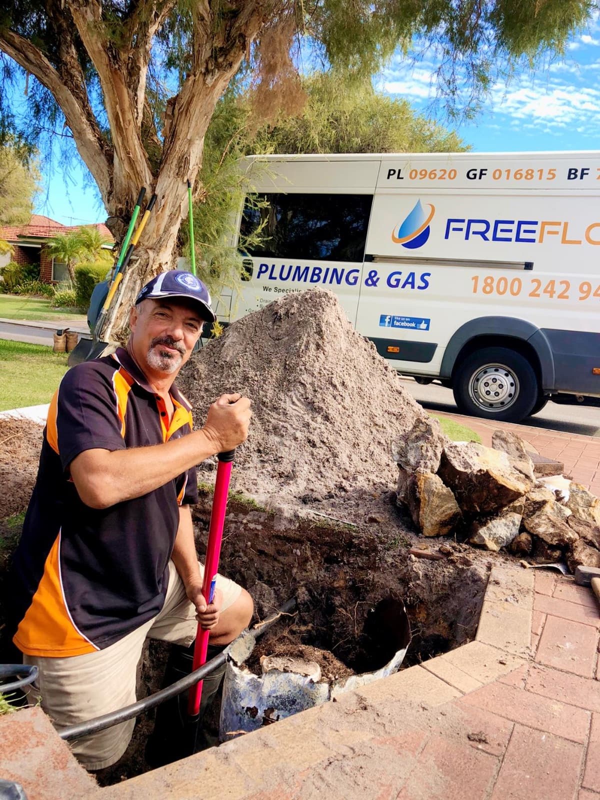 A FreeFlo plumber standing in an excavated trench in a Perth front yard, with the branded FreeFlo van behind showing its licence numbers